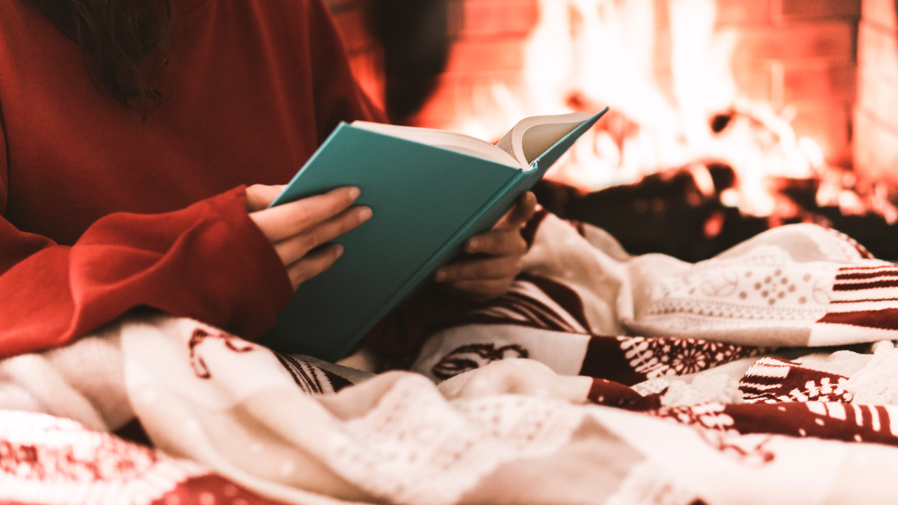 A woman reading a book as she sits next to the fireplace