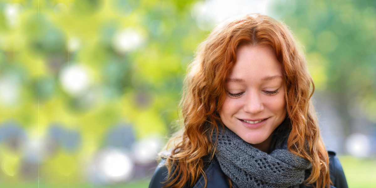 a young woman with wavy red hair looking down and smiling as she walks through a park